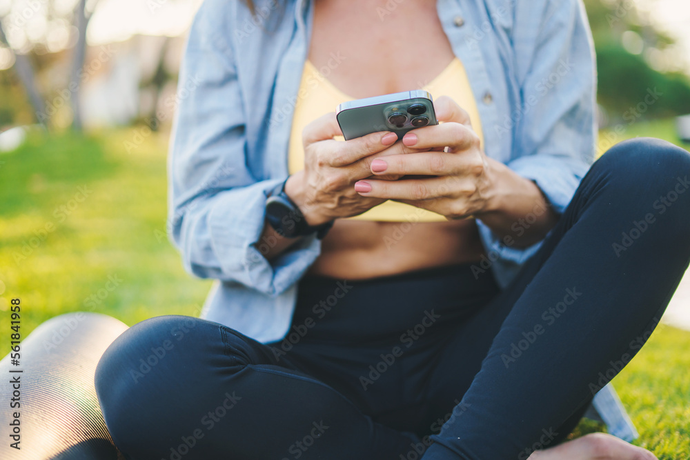 Cropped view of sporty woman chilling on green grass of athletic field and browsing mobile phone taking rest after workout. Fitness woman. Workout sport concept