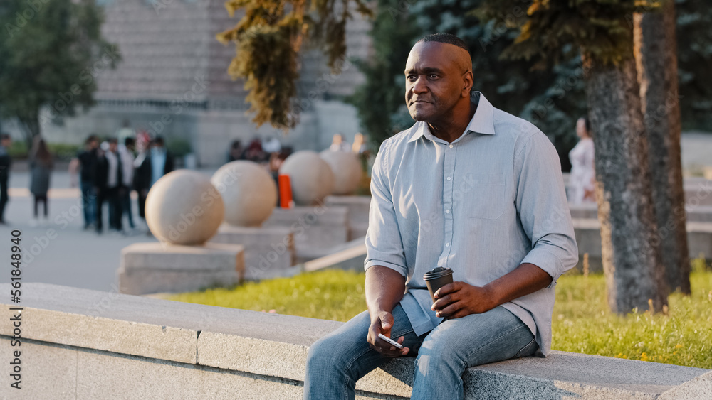 Fototapeta premium African American ethnic middle-aged man mature senior businessman male sitting outdoors in city with coffee tea and phone waiting for date business meeting looking for friend relaxing on work break