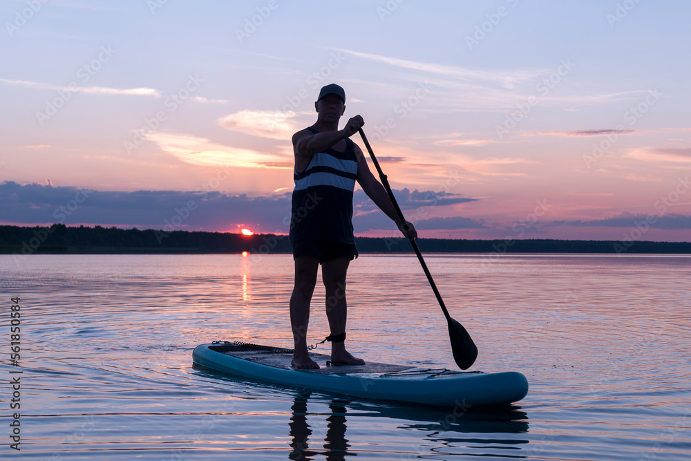A man on a SUP board with a paddle at sunset against a purple sky floats in the water of the lake.
