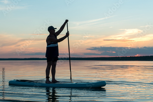 A man on a SUP board with a paddle at sunset against a purple sky floats in the water of the lake. © finist_4