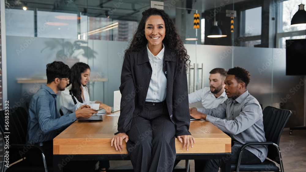 Satisfied smiling female leader boss woman company CEO businesswoman sitting on table in office smile to camera background of multiracial coworkers colleagues teamwork project startup analyze work Satisfied smiling female leader boss woman company CEO businesswoman sitting on table in office smile to camera background of multiracial coworkers colleagues teamwork project startup analyze work