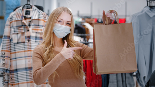 Close-up happy Caucasian woman in medical mask covid pandemic shopping stand in clothing store shop girl consumer showing pointing on bags purchases lady satisfied low prices discount Black Friday