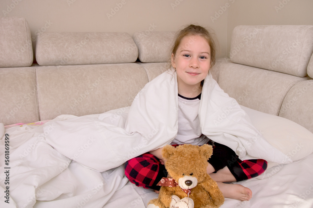 girl sits in bed in pajamas covered with a blanket with a bear and smiles