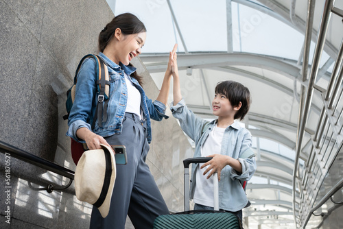 Family Trip Concept. Portrait of a cheerful Asian boy having fun and holding hands with his mother, ready for vacation, standing on the luggage cart. Parents walking with baggage enjoy a vacation.