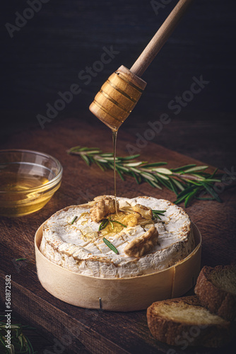 French cheese Camembert baked in a wooden box served with honey, aromatic herbs and nuts. Taste of France, rustic style. Pouring honey on cheese