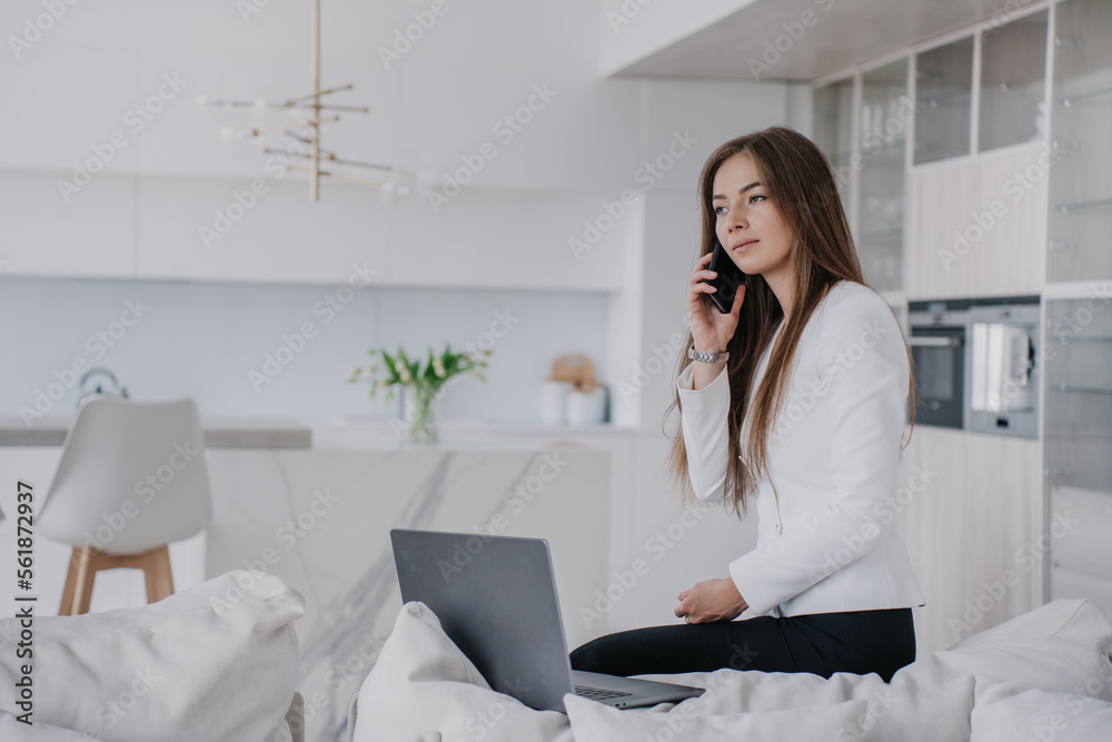 Perplexed female lawyer talking by phone with client sitting on cozy sofa dressed in business suit typing on laptop. Attractive young woman with loose hair remote working home over blurry interior. Perplexed female lawyer talking by phone with client sitting on cozy sofa dressed in business suit typing on laptop. Attractive young woman with loose hair remote working home over blurry interior.