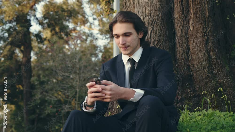Smiling businessman in the city looking to a cell phone. .Confident and happy man in a business suit is sitting outdoors and using smartphone. Young man in a suit with mobile phone, slow motion. Smiling businessman in the city looking to a cell phone. .Confident and happy man in a business suit is sitting outdoors and using smartphone. Young man in a suit with mobile phone, slow motion.