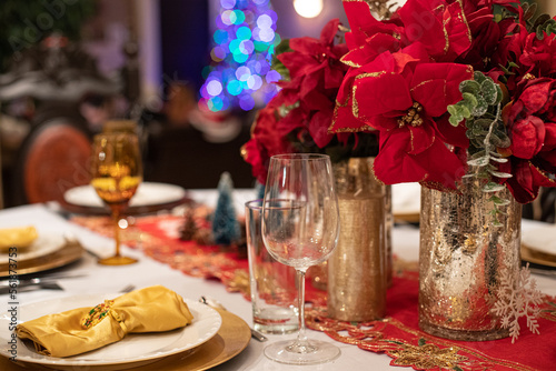 Table de fête avec couverts et verres de luxe, serviettes, assiettes et vases dorés, nappe blanche et napperon brodé rouge. Décoration et étoiles de Noël rouges.