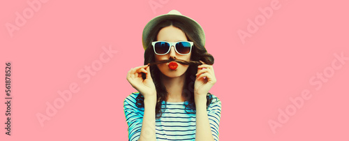 Portrait of funny young woman showing mustache her hair blowing lips with red lipstick sending air kiss on pink background © rohappy