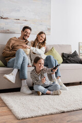 full length of positive kid clicking channels near happy parents sitting with popcorn at home.