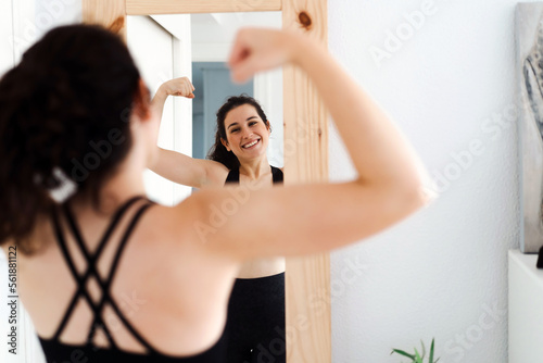 Funny young woman flexing arms looking in the mirror after training