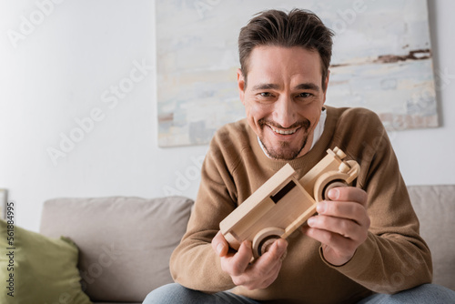 joyful man smiling while looking at camera and holding wooden car toy in living room.