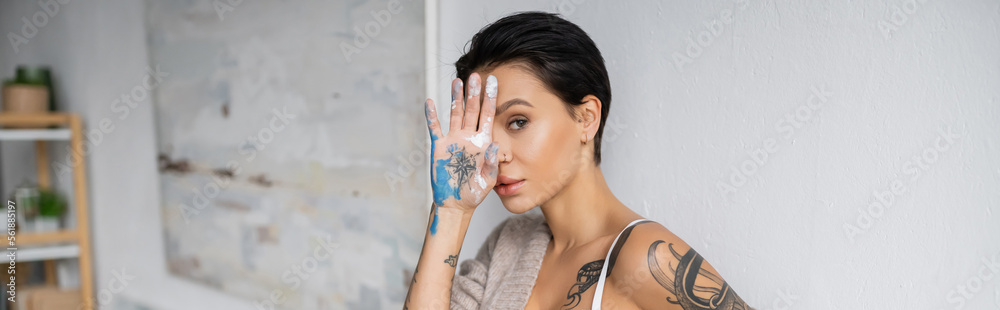 Seductive artist holding hand in paint near face and looking at camera in workshop, banner Seductive artist holding hand in paint near face and looking at camera in workshop, banner