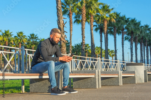 Middle aged man with slicked back hair and beard sits bench near alley of palm trees in city types message his phone.