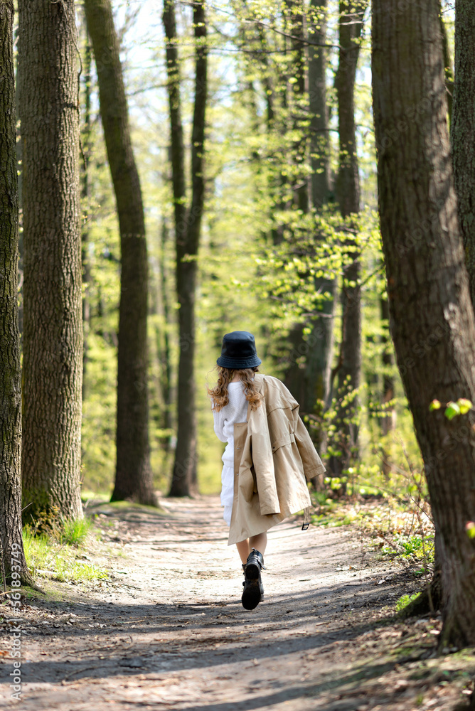 Young girl with trenchcoat and hat walks away down the road in the spring sunny forest, back view Young girl with trenchcoat and hat walks away down the road in the spring sunny forest, back view