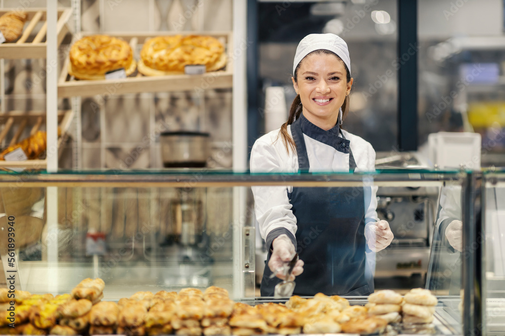 A bakery worker is selling pastry and bread at the supermarket while smiling at the camera.