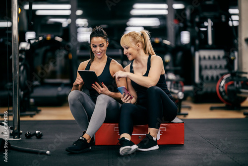 A female personal trainer is sitting next to a sportswoman and showing her progress on a tablet in a gym.