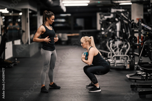 A female personal trainer is explaining how to do exercises to a sportswoman who is squatting and holding kettle bell.