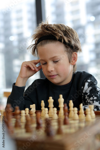 a boy of eight years, Caucasian appearance, sits in a bright room, plays chess with a friend. The boy is focused, thinks through the moves photo