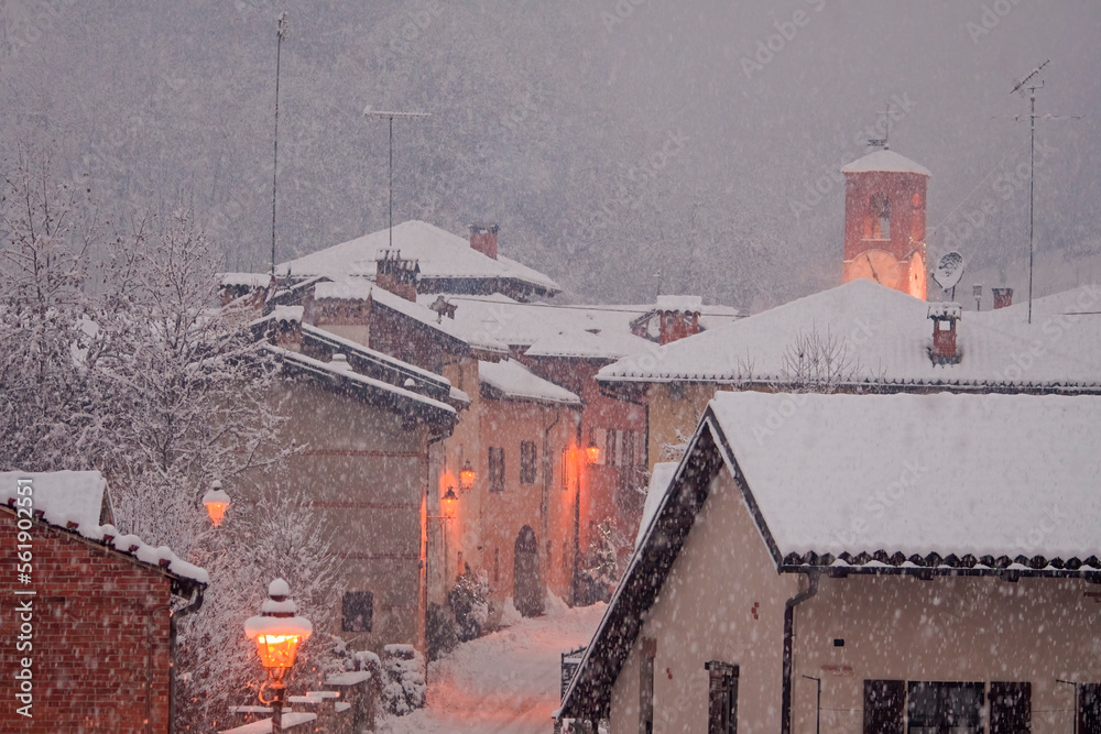 Beautiful Christmas scene of an intense snowfall in a country town with the houses and the bell tower along the central street illuminated by elegant wall lamps. Castellar, Piedmont, Italy.
