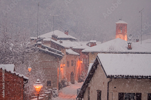 Obraz na płótnie Beautiful Christmas scene of an intense snowfall in a country town with the houses and the bell tower along the central street illuminated by elegant wall lamps. Castellar, Piedmont, Italy. w salonie