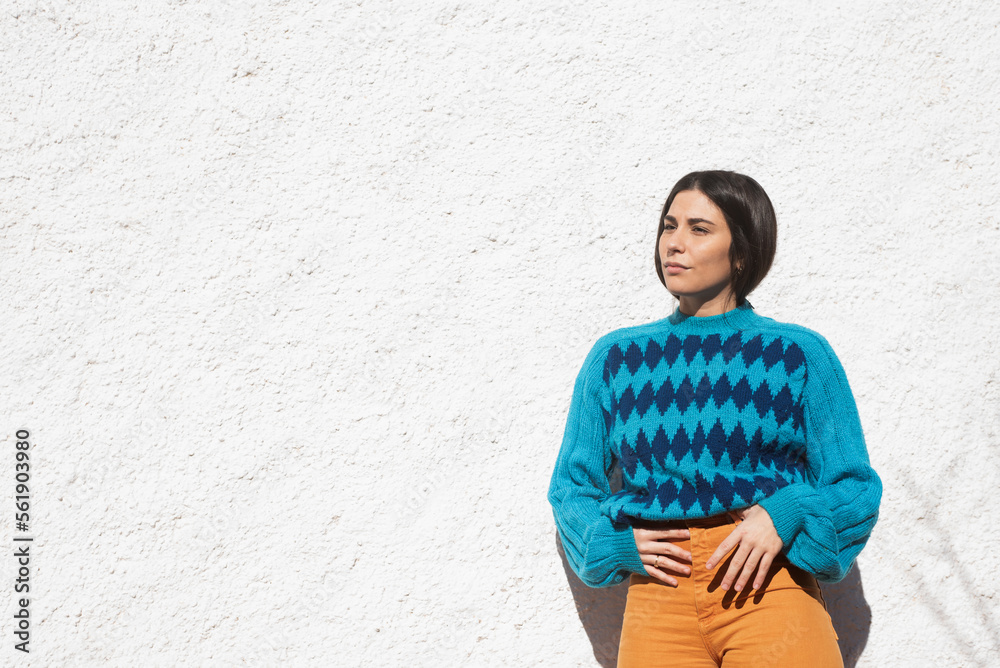 young latin girl in colorful clothes, saturated blue argyle sweater and orange pants, white stucco wall background outside, light sunny day. Copy space on one side