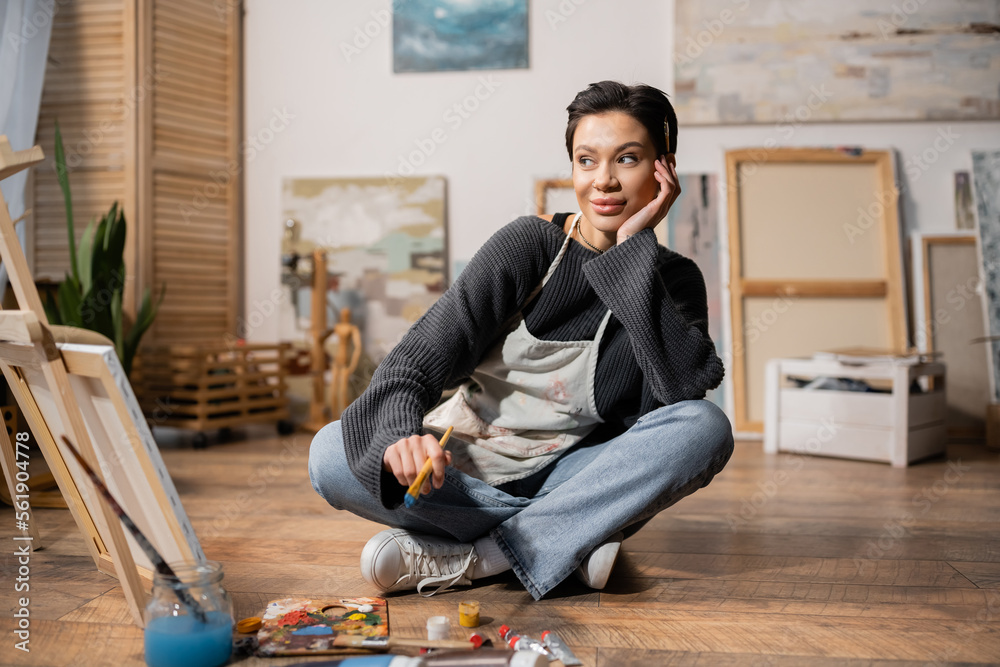 Fototapeta premium Positive artist in apron holding paintbrush near canvas and easel in workshop