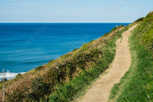 A trail path runs along the steep coast, with the sea in the background on a sunny day.