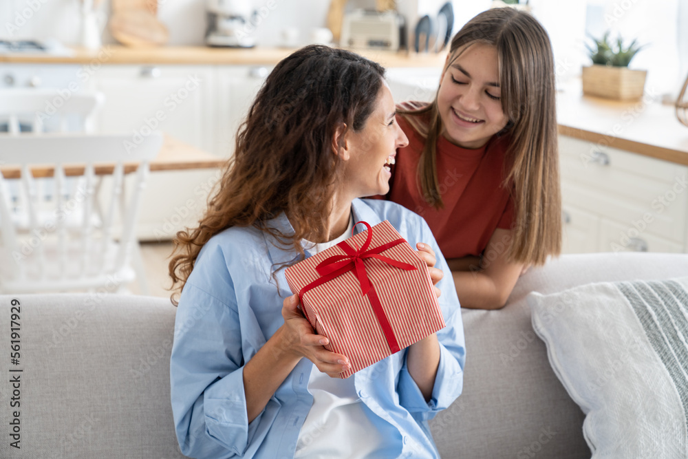 Cute teen girl loving daughter congratulating happy young mother with birthday at home, give her gift box with red ribbon. Child surprising mom on Mothers Day, kid making mum happy. Family holidays Cute teen girl loving daughter congratulating happy young mother with birthday at home, give her gift box with red ribbon. Child surprising mom on Mothers Day, kid making mum happy. Family holidays