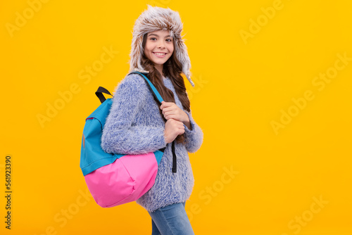 happy child in sweater and earflap hat with backpack on yellow background with copy space, school