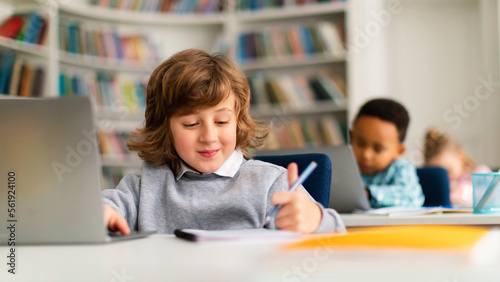 Caucasian boy studying with laptop and writing in notepad, sitting at table in classrom with diverse classmates photo