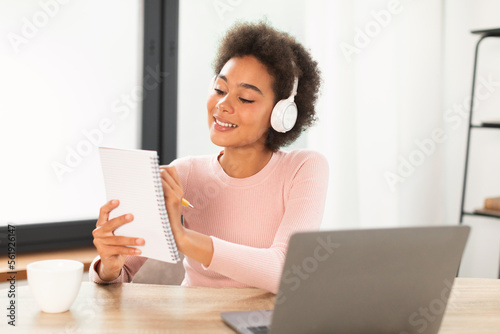 Smiling dreamy pensive young mixed race female manager in wireless headphones makes notes