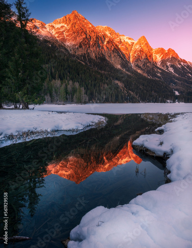 Snowcapped Alps mountains reflected with warm colors in the partially frozen Anterselva lake (North Italy) at sunset in winter. Snow and ice in the foreground, tree and forest in the background.