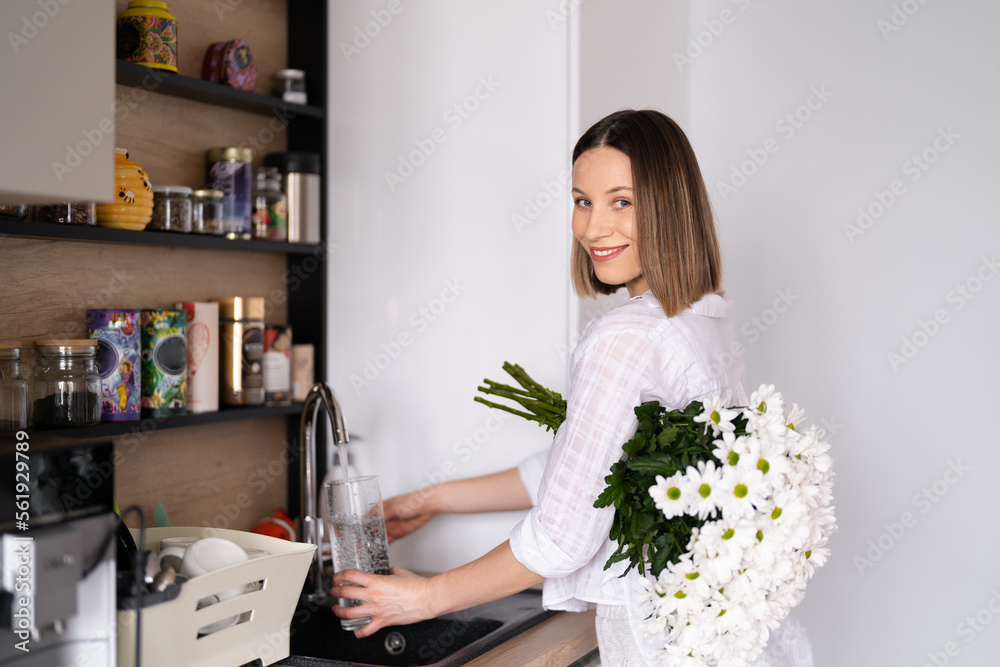 Positive pretty young woman in white casual clothes holding a bunch of white flowers while pouring water into vase in domestic kitchen Positive pretty young woman in white casual clothes holding a bunch of white flowers while pouring water into vase in domestic kitchen