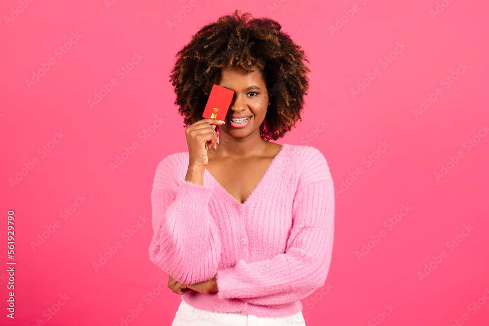 Happy millennial african american curly woman shopaholic in casual puts credit card to eye, has fun Happy millennial african american curly woman shopaholic in casual puts credit card to eye, has fun