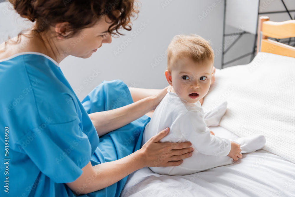 baby girl with open mouth looking away while sitting near mom on bed in clinic. baby girl with open mouth looking away while sitting near mom on bed in clinic.