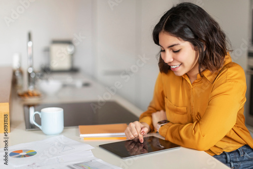 Smiling Young Arab Female Browsing Internet On Digital Tablet In Kitchen