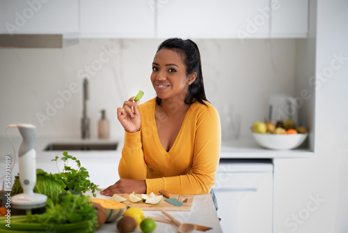 Smiling young african american female eating piece of fruit at table with organic vegetables