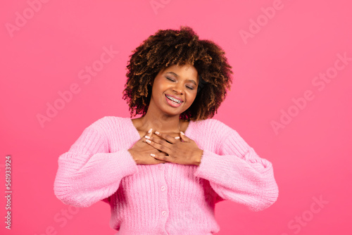 Smiling pretty millennial african american curly female in casual wear presses hands to chest, enjoy thankful