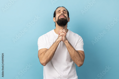 Man wearing white T-shirt looking up holding hands together, praying God, asking for health and support pleading for healing and forgiveness. Indoor studio shot isolated on blue background.
