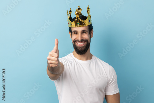 Photography Portrait of smiling joyful man with beard wearing white T-shirt and in gold crown standing looking at camera with toothy smile, showing thumb up