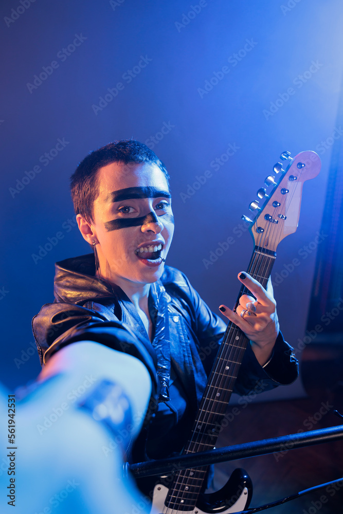 Obraz premium Punk rocker showing rock sign on camera and playing guitar, creating musical performance and acting crazy. Artist producing heavy metal alternative music with instrument, live performance.