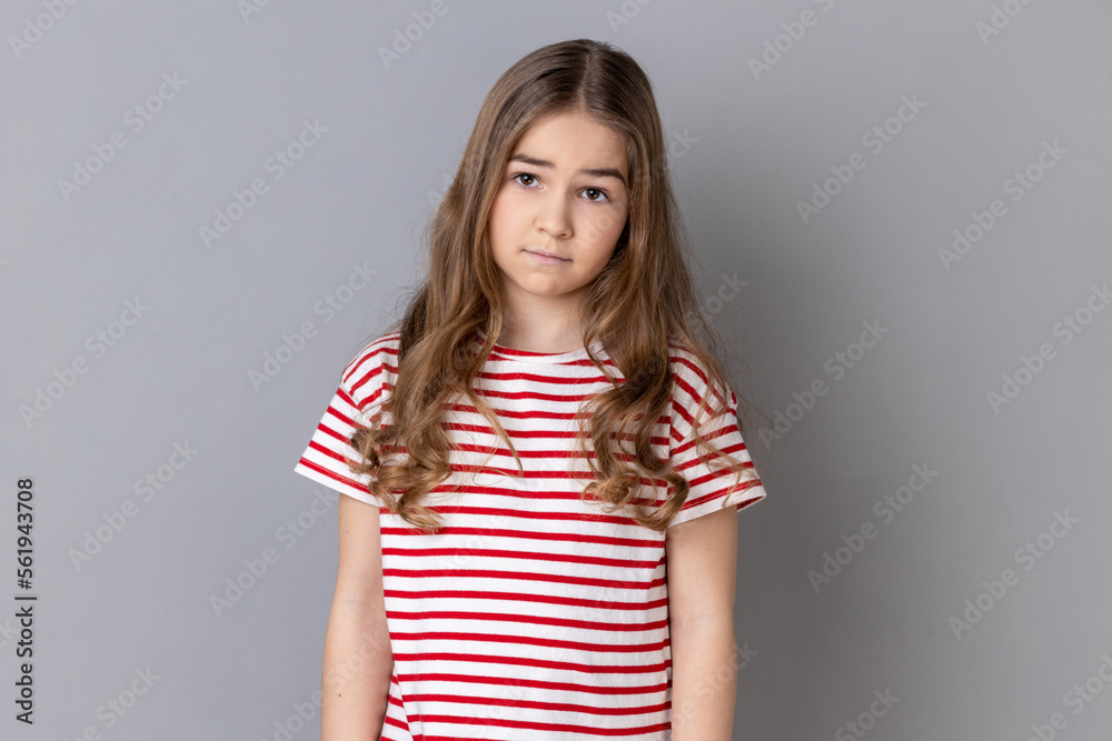 Portrait of unhappy little girl wearing striped T-shirt being upset of bad news, looking at camera with frowning face, expressing sadness. Indoor studio shot isolated on gray background. Portrait of unhappy little girl wearing striped T-shirt being upset of bad news, looking at camera with frowning face, expressing sadness. Indoor studio shot isolated on gray background.