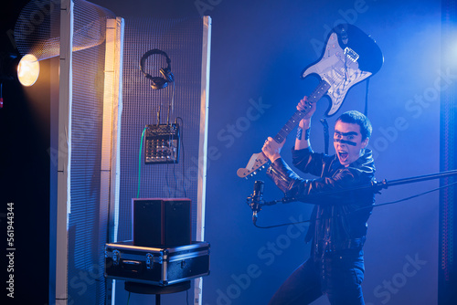 Ecstatic artist holding bass guitar to smash and act crazy, performing punk rock music and screaming loud in studio. Rocker with leather jacket fooling around and throwing musical instrument.