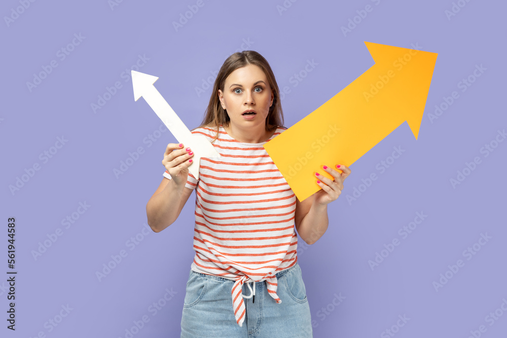 Portrait of shocked scared blond woman wearing striped T-shirt holding two arrows indicating to different sizes, looking at camera with big eyes. Indoor studio shot isolated on purple background. Portrait of shocked scared blond woman wearing striped T-shirt holding two arrows indicating to different sizes, looking at camera with big eyes. Indoor studio shot isolated on purple background.