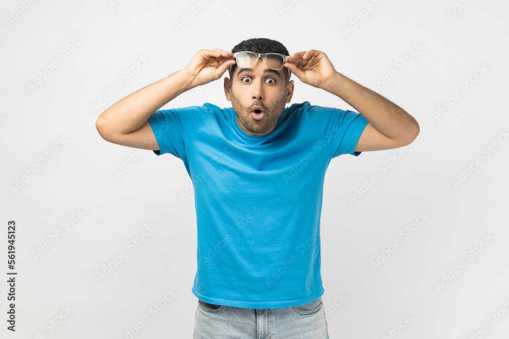 Portrait of shocked astonished unshaven man wearing blue T- shirt standing raised his optical eyeglasses, sees something surprised. Indoor studio shot isolated on gray background. Portrait of shocked astonished unshaven man wearing blue T- shirt standing raised his optical eyeglasses, sees something surprised. Indoor studio shot isolated on gray background.