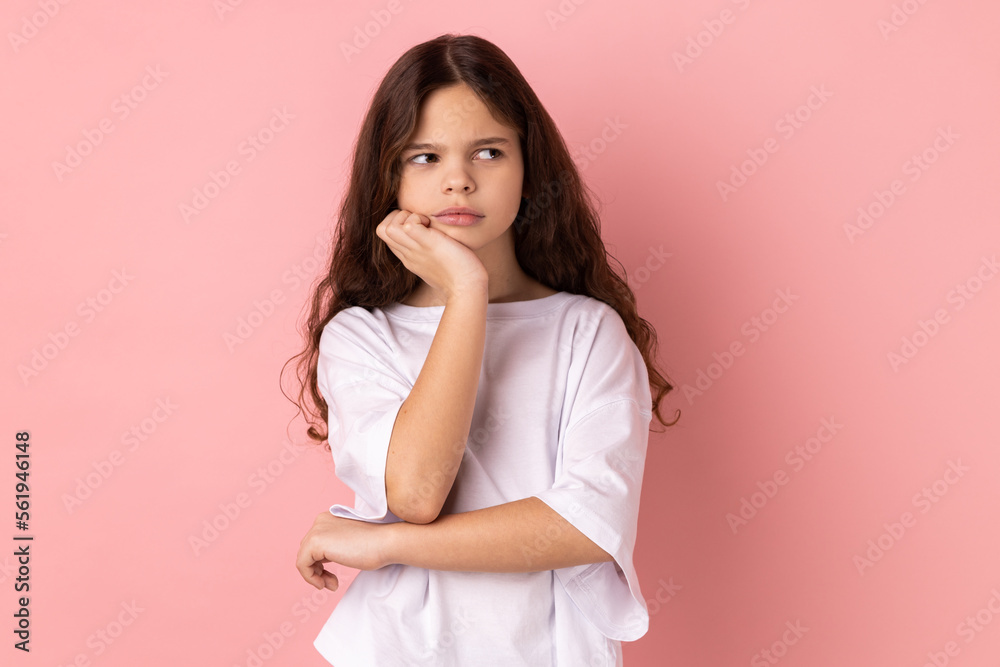Portrait of thoughtful pensive little girl wearing white T-shirt thinking about future, holding chin, having serious facial expression. Indoor studio shot isolated on pink background.