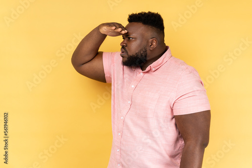 Side view of curious man with beard in pink shirt keeping palm over head and looking attentively far away, peering with expectation at long distance. Indoor studio shot isolated on yellow background. photo