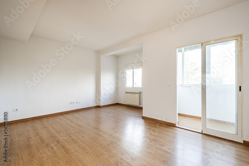 Empty room with a light wooden floor, a white aluminum radiator under a window and a closed terrace with a white aluminum and glass sliding door