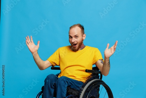 a man sits in a wheelchair thoughtfulness in a T-shirt on a blue background in the studio, Raised his hands up and opened his mouth in surprise, tattooed body